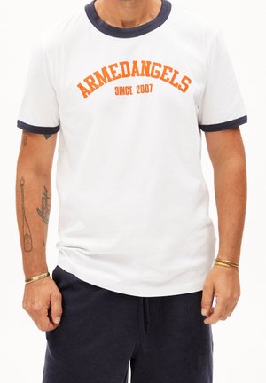 Homme portant un T-shirt blanc avec une bordure noire et un texte orange "ARMEDANGELS SINCE 2007,", pantalon noir, bracelets et tatouages visibles sur les bras.