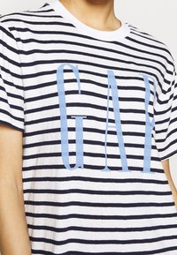 Pasiasta bawełniana koszulka z niebieskimi i białymi poziomymi paskami, z dużym, jasnoniebieskim logo "GAP" na środku. Okrągły dekolt.