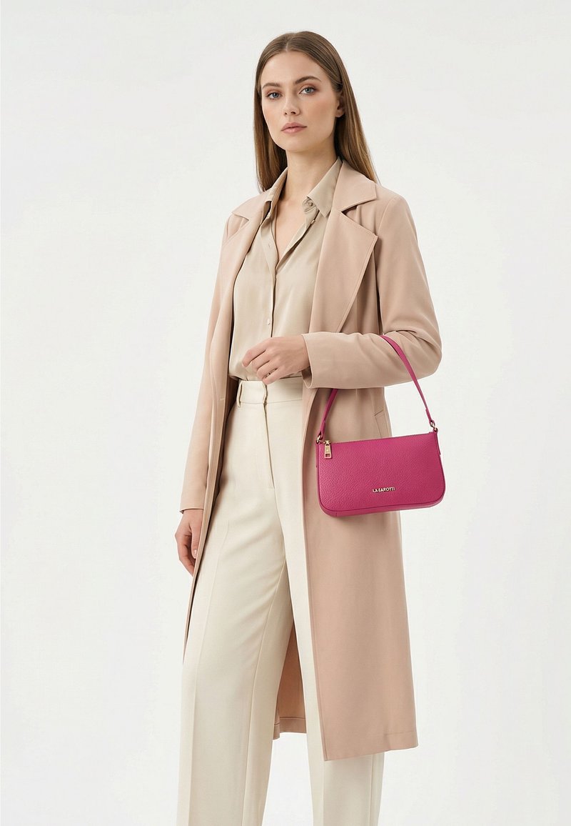 Mujer con abrigo beige, blusa crema y pantalones, sosteniendo un pequeño bolso de mano de color rosa brillante en su brazo izquierdo contra un fondo liso y claro.