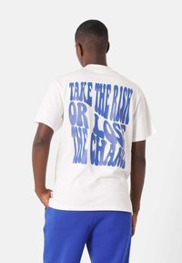 Hvid bomulds t-shirt med stor blå tekst på ryggen: "TAG RISIKOEN ELLER TAB MULIGHEDEN." Korte ærmer, afslappet pasform, ensfarvet.
