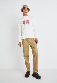 White sweatshirt with a graphic print, beige pants, and dark shoes with yellow laces. Complemented by a beige bucket hat.