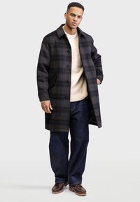 Karierten Mantel aus einem dunklen grauen und schwarzen Wollgemisch, mit Knopfleiste, Kragen und Seitentaschen, getragen über einem cremefarbenen Pullover und blauen Jeans.