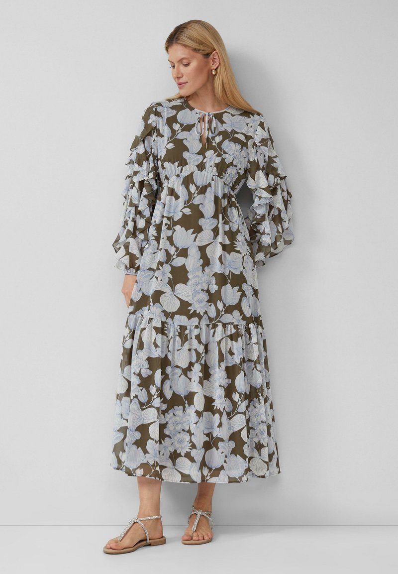 Maxikleid aus olivgrünem Stoff mit hellblauen Blumen, mit gebundenem Hals, rüschenslangen Ärmeln und gestuftem Rock. Getragen mit flachen Sandalen.
