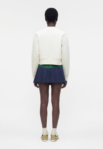 Άτομο που στέκεται με τα πίσω στραμμένα, φοράει κοντό λευκό φούτερ, πράσινο πουκάμισο, σκούρη μπλε πλισέ φούστα, λευκές κάλτσες και μπεζ αθλητικά παπούτσια σε λευκό φόντο.