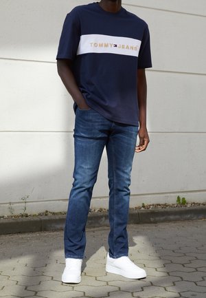 Man met een navyblauw Tommy Jeans T-shirt, blauwe spijkerbroek en witte sneakers, staand buiten op een bestrate ondergrond bij een witte muur.