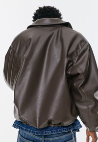 Hnedá syntetická kožená bunda s voľným strihom, výraznými ramennými švami a zaobleným golierom. Textília má jemnú štruktúru.