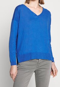 Blaues V-Ausschnitt-Pullover aus leichtem Strickstoff mit langen Ärmeln und seitlichen Schlitzen, kombiniert mit hellgrauen, schmal geschnittenen Jeans.