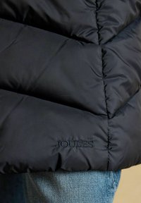 Melnā izšūta jaka ar 'JOULES' uzrakstu tuvu apakšai, valkāta virs gaiši zilām džinsa biksēm.