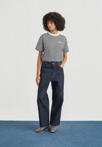 Γυναίκα με σγουρά μαλλιά, ντυμένη με ριγέ πουκάμισο σε μαύρο και άσπρο, σκούρα τζιν με φαρδιά μπατζάκια και γκρι αθλητικά, στέκεται σε μπλε χαλί.