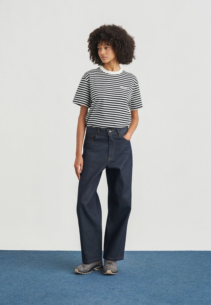 Kvinde med krøllet hår iført en sort-hvid stribet skjorte, mørke vide jeans og grå sneakers, stående på blåt tæppe.