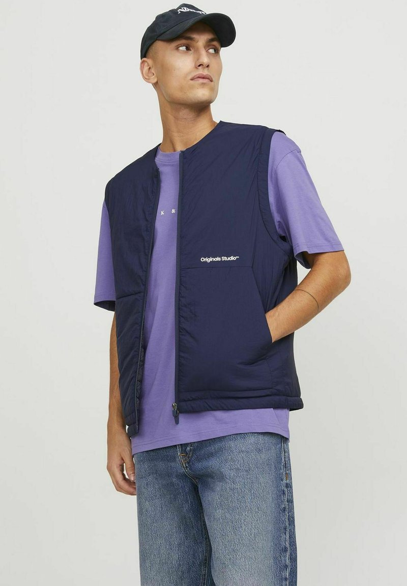Gilet matelassé bleu marine avec fermeture éclair, arborant une poche poitrine et un logo. Porté sur un t-shirt violet clair et associé à un jean bleu.