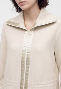 Femme portant une veste beige avec fermeture éclair, un large col côtelé et une bordure en satin brillante le long de la fermeture éclair et des bords du col.