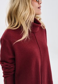 Czerwony sweter z golfem o miękkiej fakturze, charakteryzujący się luźnym krojem i rolowanymi mankietami. Zauważalne detale szwów na froncie i po bokach.