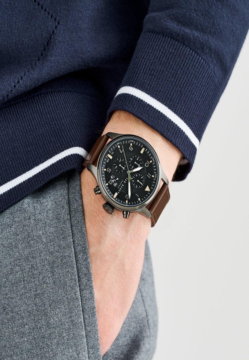 Fekete kronográf óra barna bőrszíjjal, ezüst részletekkel és texturált számlappal. Szürke szövet hátterű csuklón viselve.