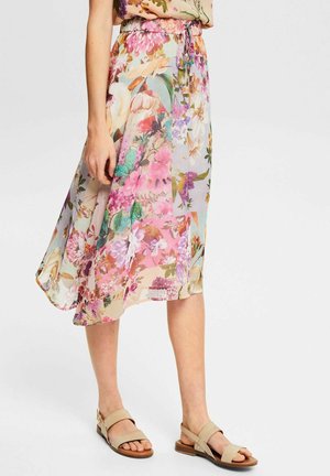 Femme portant une jupe mi-longue transparente avec des motifs floraux roses, violets et verts, associée à des sandales beiges ouvertes aux orteils.