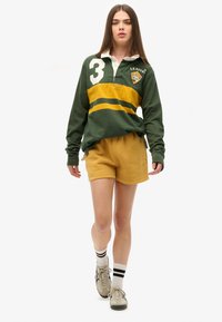 Femme marchant avec un maillot de rugby vert et jaune portant le numéro 3, un short moutarde, des chaussettes blanches à rayures noires et des baskets beiges.