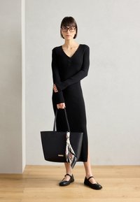 Vestido de punto acanalado negro con escote en V y detalles de botones, combinado con un bolso tote negro adornado con una bufanda floral y unas bailarinas negras.