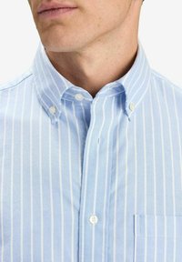 Vaaleansininen nappikauluspaita, jossa on ohuita valkoisia raitoja, kaulus ja rintatasku. Varustettu muovipainikkeilla ja tekstiilitulella.