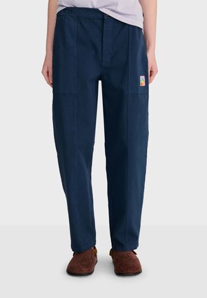 Personne portant un pantalon droit bleu marine avec une petite pièce colorée sur la partie supérieure de la cuisse et des chaussons marron à l'intérieur sur un fond uni.
