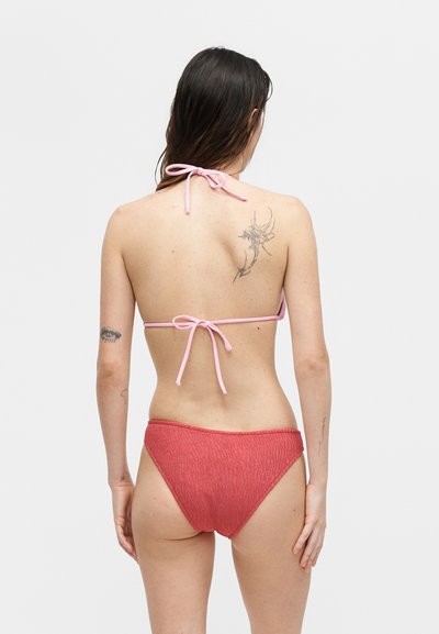 Γυναίκα με μακριά καστανά μαλλιά, που φορεί ένα ροζ μπικίνι τοπ και κόκκινο υφασμάτινο μπικίνι κάτω μέρος, γυρισμένη με την πλάτη, δείχνοντας τα τατουάζ στην πλάτη και τα χέρια.