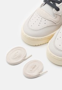 Zapatillas gris claro con un acabado de cuero suave, puntera perforada y suela de goma beige. Se incluyen dos cordones beige al lado de los zapatos.