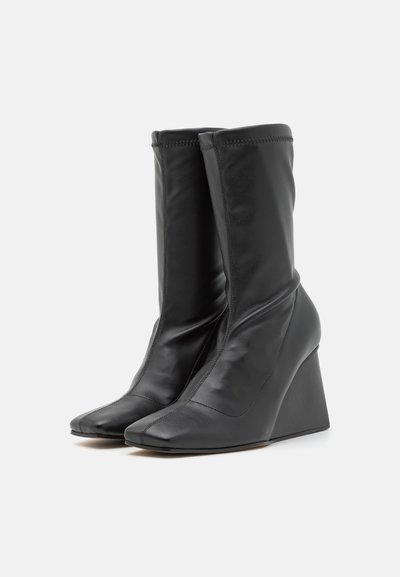 Cizme negre din piele cu toc wedge, la gleznă, cu un vârf ascuțit și textură netedă. Prezintă un șiret ajustat și detalii de cusătură minimă.