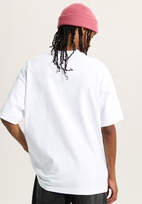 Camiseta blanca de algodón con corte relajado, mangas cortas y escote redondo, combinada con un gorro de lana rosa. Se muestra la vista trasera.