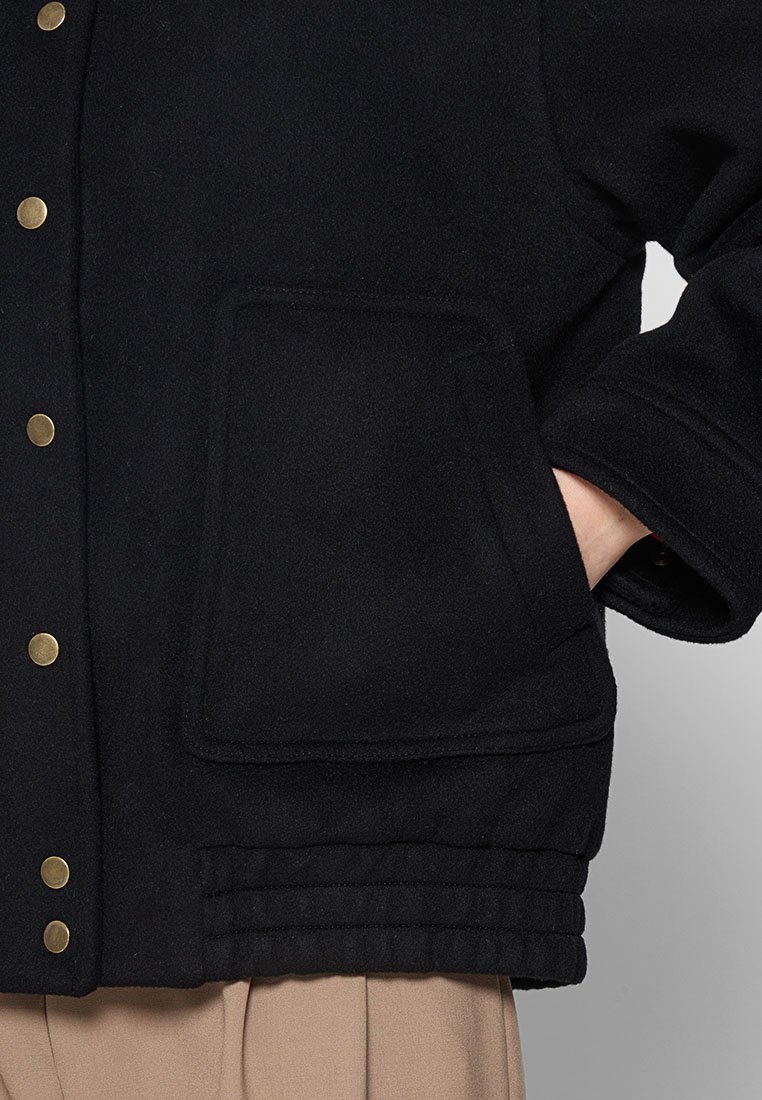 Schwarze Fleecejacke mit einer Fronttasche, goldenen Druckknöpfen und strukturierten Rippdetails am Saum. Einfaches Design, lässige Passform.