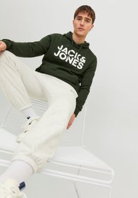 Dark green hoodie with white "JACK & JONES" logo, paired with light grey sweatpants. Model seated on a white chair, wearing white sneakers.