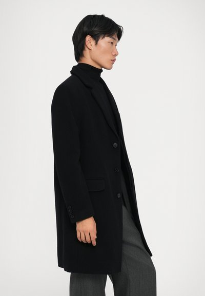 Cappotto nero con un design su misura, bottoni a un solo petto, rever a punta e tasche laterali, indossato sopra un dolcevita nero e pantaloni grigi.