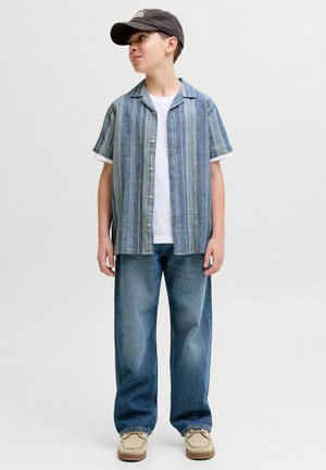 Berniukas, dėvintis juodą kepurę, mėlyną dryžuotą trumpomis rankovėmis marškinių virš baltos maikės, laisvus mėlynus džinsus ir smėlio spalvos batus, stovintis ir žiūrintis į šoną.