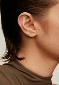 Pendiente de plata con una pequeña gema cuadrada transparente, visible en la oreja de un modelo. El cabello es oscuro y de longitud hasta los hombros, parcialmente visible.