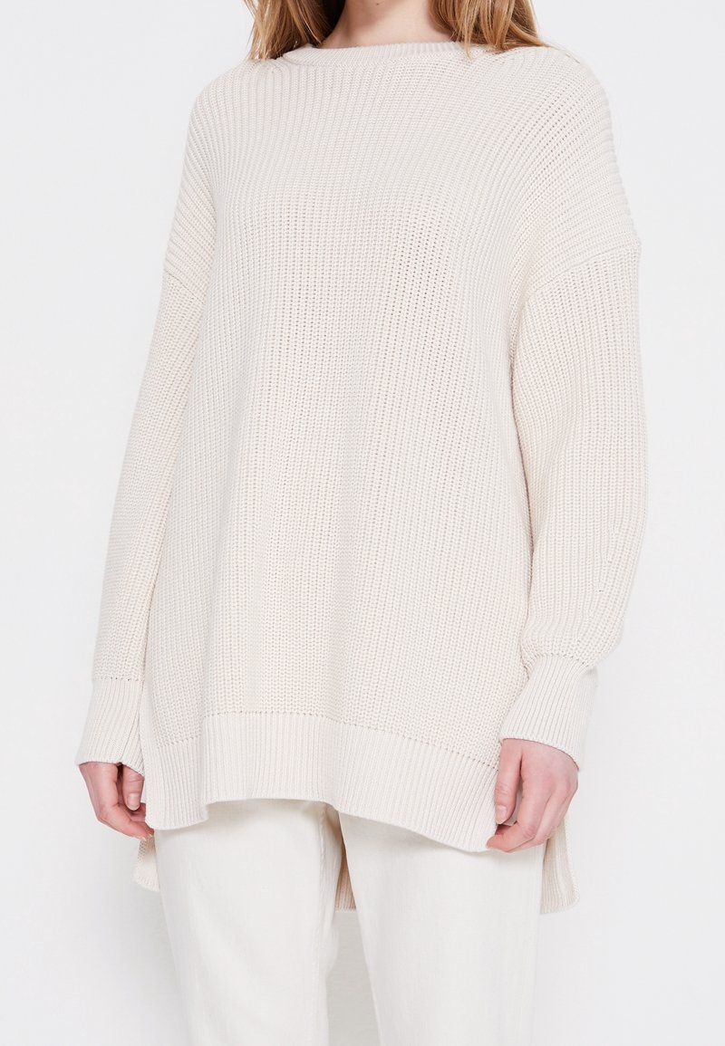 Žena v přes oversized krémovém pleteném svetru s vykrojenými rameny a asymetrickým lemem, stojící proti bílému pozadí, v bílé kalhoty.