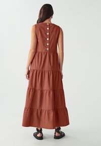 Maxikleid in Rostfarbe mit lässiger Passform, gestuftem Design, runder Ausschnitt und Knopfdetails auf der Rückseite. Aus weichem Stoff und ärmellos.