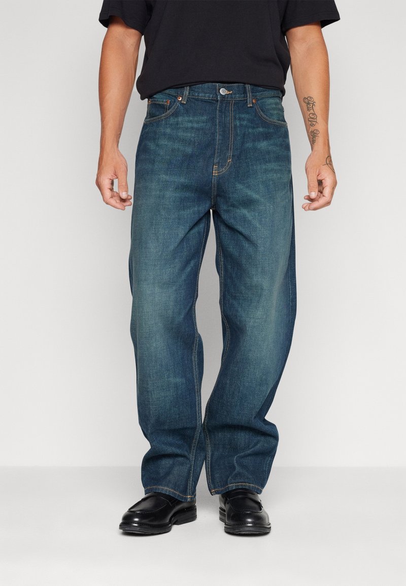 Homme portant un jean bleu ample, des chaussures noires et une chemise noire à manches courtes, debout devant un fond clair uni.