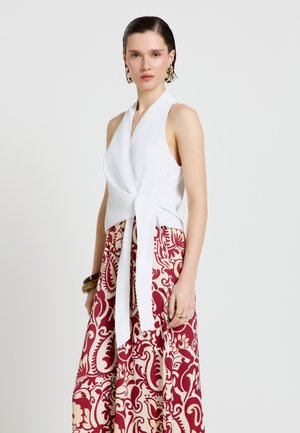 Frau mit zurückgestrichenen Haaren, die ein ärmelloses weißes Wickeltop und rote gemusterte weite Hosen trägt, ergänzt mit goldenen Ohrringen und Armbändern.