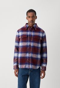 Red, blue, and white plaid flannel shirt with button-up front, two chest pockets, and a collar; features textured fabric and relaxed fit.