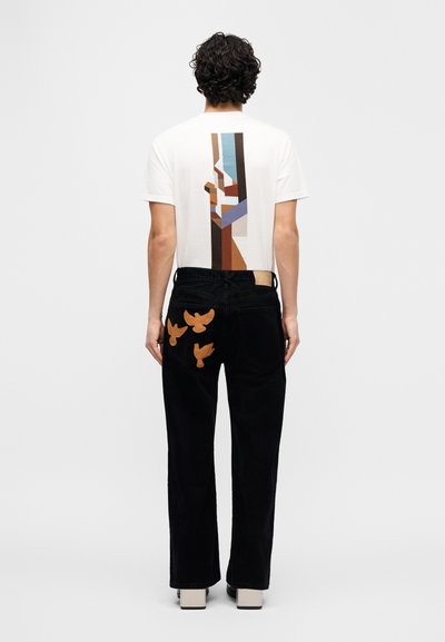 Λευκό μπλουζάκι με γεωμετρικό σχέδιο σε αποχρώσεις καφέ, μπλε και ταμπά, συνδυασμένο με μαύρο παντελόνι από κορντόρι με καφέ κεντήματα πουλιών.