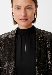 Czarna marynarka z cekinami o gładkiej strukturze, z wysokim marszczonym dekoltem i otworem w kształcie łezki. Uzupełniona dużymi srebrnymi kolczykami.
