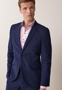 Blazer azul marino con dos botones, solapas con muesca y un bolsillo en el pecho. Llevado sobre una camisa rosa clara con un cuello decorativo y botones oscuros.