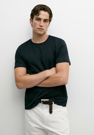 Junger Mann mit verschränkten Armen, trägt ein schwarzes T-Shirt und weiße Hosen mit einem geflochtenen schwarzen Gürtel, steht vor einer weißen Wand.