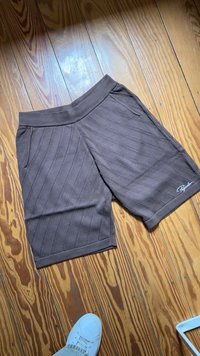 Pantaloni corti marroni realizzati in tessuto texturizzato con strisce diagonali, tasche laterali e una vita a coste. Logo del marchio esposto in blu.