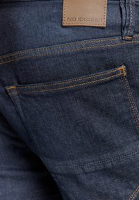 Donkerblauwe denim jeans met oranje stiksels, voorzien van een leren patch met de tekst "NO EXCESS" op de achterkant van de taille. Inclusief achterzakken.