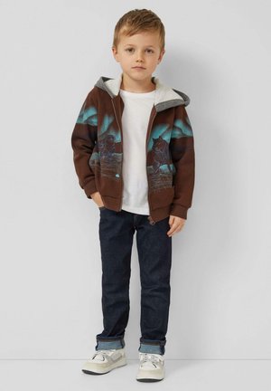 Sweat à capuche marron zippé avec doublure grise, orné d'un motif graphique bleu, t-shirt blanc, jean bleu foncé, et baskets claires.