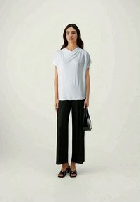 Bílý top s látkovým přehozem v kombinaci s černými širokými plisovanými kalhotami, černými sandály na otevřenou špičku a černou kabelkou, na neutrálním pozadí.