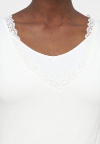 Άτομο με σκούρο δέρμα που φοράει λευκό πουκάμισο με λαιμόκοψη σε σχήμα V διακοσμημένη με λεπτή λευκή δαντέλα.