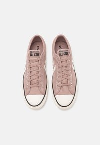 Jasnoróżowe zamszowe sneakersy mają białą gumową osłonę palców, czarne wykończenia oraz klasyczny design z wiązaniem, srebrnymi oczkami oraz brandingiem na języku.