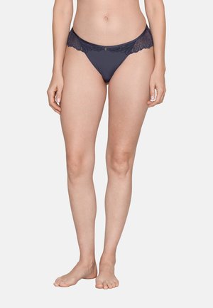 Weiblicher Torso und Beine, die dunkelblaue Spitzenbikiniunterwäsche tragen, stehen barfuß auf einem schlichten hellen Hintergrund.