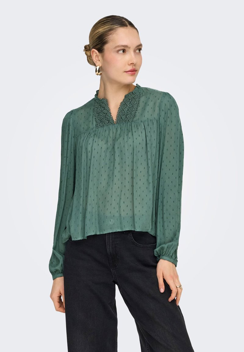 Γυναίκα με μαλλιά σε κότσο, που φορεί πράσινο πουκάμισο με μοτίβο και λεπτομέρειες από δαντέλα στα μανίκια, συνδυασμένο με μαύρο παντελόνι, κοιτάζει προς τα πλάγια.