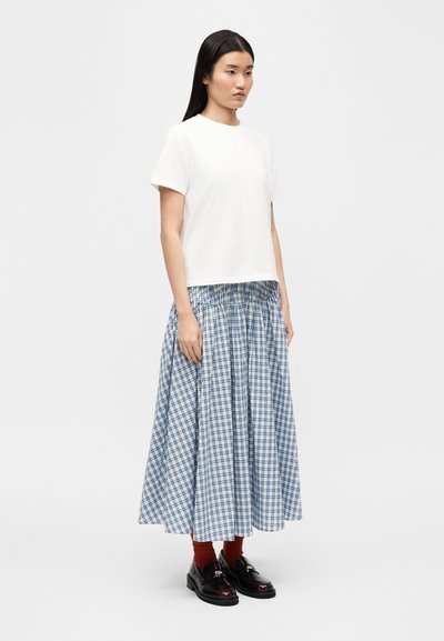 Femme debout, portant une chemise blanche à manches courtes, une jupe midi à carreaux bleus et blancs, des chaussettes couleur rouille et des mocassins noirs sur un fond uni.